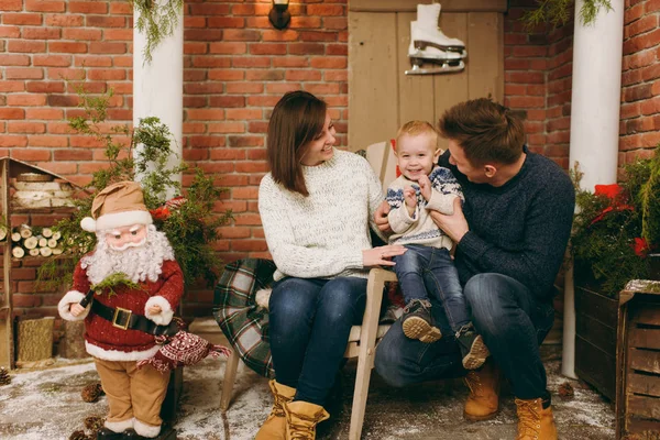 Young happy parents with a cute little child boy dressed in sweater sitting on the chair with Santa in decorated New Year room at home. Christmas good mood. Lifestyle, family and holiday 2018 concept