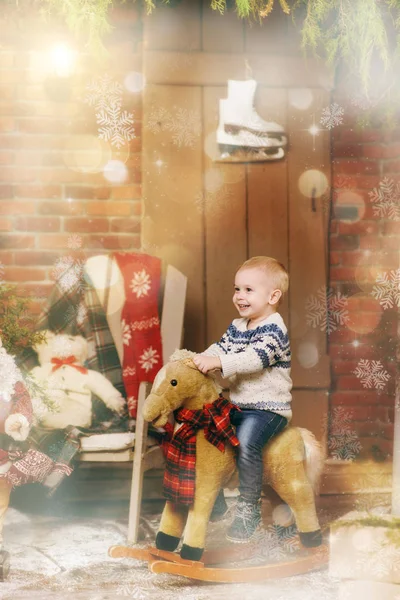 Juguetón sonriente niño feliz sentado en el caballo mecedora en la habitación decorada de Año Nuevo en casa. Navidad, hijo buen humor. Estilo de vida, familia y vacaciones 2018 concepto. Resalte mágico efecto bokeh — Foto de Stock