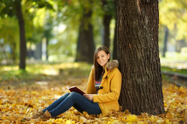 A beautiful happy smiling brown-haired woman in yellow coat and jeans sitting under the maple tree with a red book in fall city park on a warm day. Autumn golden leaves. Reading concept — Stock Photo, Image