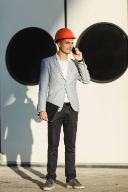 Gri takım elbise ve koruyucu inşaat turuncu kask, yakışıklı genç tıraşsız başarılı iş adamı cep telefonda konuşurken liman yuvarlak windows arka plan kapılarla karşı ayakta