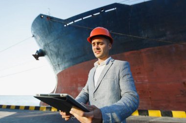 Yakışıklı tıraşsız başarılı iş adamı gri takım elbise, tablet, liman kargo paslı geminin su hattı arka plan üzerinde karşı ayakta tutan koruyucu inşaat turuncu kask. Erkek ile becerikli alet