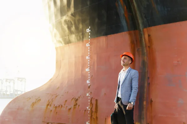 Handsome young unshaven successful business man in gray suit and protective construction orange helmet holding case, standing in sea port against a cargo rusty ship with water line background