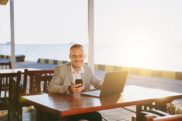 Красивый небритый молодой бизнесмен в костюме с мобильным телефоном, читающий смс, делающий заметки и улыбающийся во время работы за столом с ноутбуком в солнечном кафе. Концепция фриланса . — стоковое фото