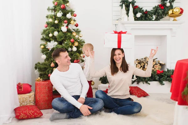 Jovens pais alegres felizes com o filhinho bonito. Menino sentado na sala de luz em casa com árvore de Ano Novo decorada e caixa de presente na cabeça. Bom humor de Natal. Conceito de família, amor e férias 2018 — Fotografia de Stock