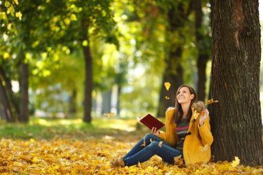 A beautiful happy smiling brown-haired woman in yellow coat and jeans sitting under the maple tree with a red book in fall city park on a warm day. Autumn golden leaves. Reading concept clipart
