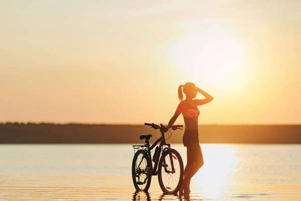 Silna Kobieta blonde w kolorowy komplet stoi w pobliżu rowerów w wodzie o zachodzie słońca na ciepły, letni dzień. Koncepcja fitness. Tło nieba — Zdjęcie stockowe