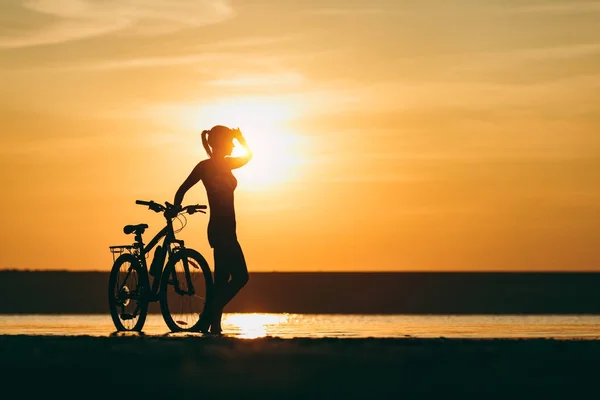 Sylwetka dziewczynki sportowy w garniturze stojącego w pobliżu rower w wodzie o zachodzie słońca na ciepły, letni dzień. Koncepcja fitness. Tło nieba — Zdjęcie stockowe