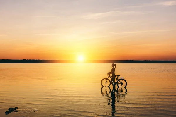 Silhouette di una ragazza sportiva in un abito che si siede su una bicicletta in acqua al tramonto in una calda giornata estiva. Concetto fitness. Sfondo cielo — Foto Stock