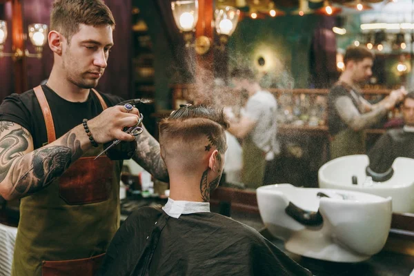 Чоловік стає модною стрижкою в перукарні. Чоловічий перукар в татуюваннях, що обслуговують клієнта . — стокове фото