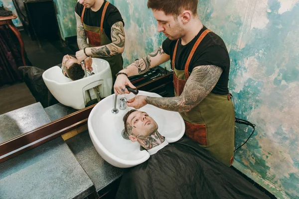 Um homem a cortar o cabelo na barbearia. Cabeleireiro masculino em tatuagens que servem cliente, cabeça de lavagem — Fotografia de Stock