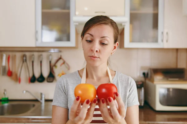 Μια νεαρή γυναίκα ελκυστική σκεπτικός σε μια ποδιά αποφασίζει να επιλέξουν μια κόκκινη ή κίτρινη ντομάτα στην κουζίνα. Δίαιτα έννοια. Υγιεινού τρόπου ζωής. Το μαγείρεμα στο σπίτι. Προετοιμασία φαγητού. — Φωτογραφία Αρχείου