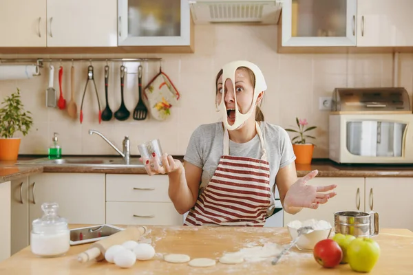 Μια νεαρή αστεία, χαρούμενη και χαμογελαστή γυναίκα βάζει σε μια ζύμη με τρύπες στο πρόσωπό της και έχει τη διασκέδαση στην κουζίνα. Μαγείρεμα σπίτι. Προετοιμασία φαγητού. — Φωτογραφία Αρχείου