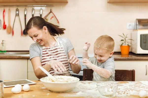 Niño pequeño ayuda a la madre a cocinar galletas de jengibre de Navidad en la cocina ligera con la tableta en la mesa. Feliz familia mamá 30-35 años y el niño 2-3 divertirse y tirar harina a casa. Concepto de relación —  Fotos de Stock