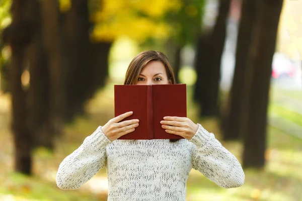 A beautiful happy smiling brown-haired woman in white sweater hiding behind a red book in fall city park on a warm day. Autumn golden leaves. Reading concept.