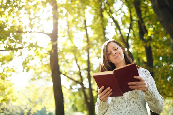 Güzel bir mutlu gülümseyen kahverengi saçlı kadın beyaz kazak daimi sıcak bir günde sonbahar Şehir Parkı içinde kırmızı bir kitapla. Altın yaprak. Okuma kavramı.