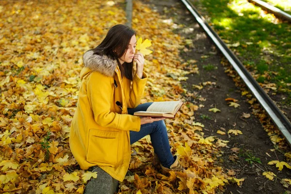 Piękne kobiety Brunet szczęśliwy w żółtym fartuchu i dżinsy samodzielnie znajduje się w parku w pobliżu torów tramwajowych i czyta książkę w ciepły dzień jesieni. Żółty jesienny liść. — Zdjęcie stockowe