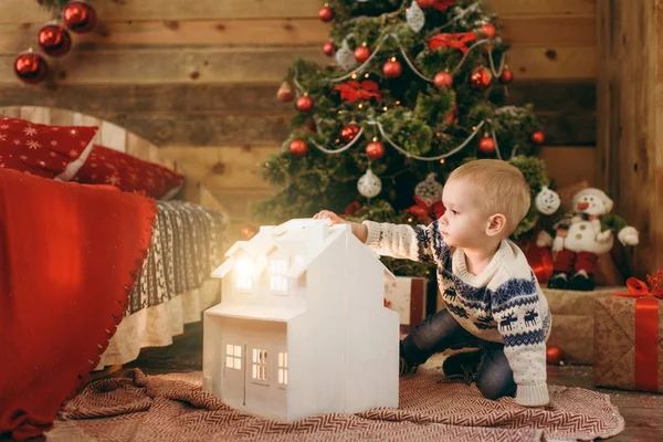 Feliz niño lindo vestido con suéter sentado en el árbol de Navidad con una casa blanca de juguete mágico en la habitación de madera en casa. Buen humor para niños. Año Nuevo. Estilo de vida, familia y vacaciones 2018 concepto — Foto de Stock