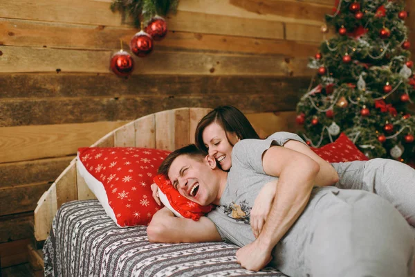 Coppia amorevole in pigiama grigio appoggiato sul letto vicino all'albero di Natale in camera decorata Capodanno a casa. Abbraccia l'atmosfera natalizia e il comfort. Stile di vita, famiglia e vacanza 2018 concept — Foto Stock