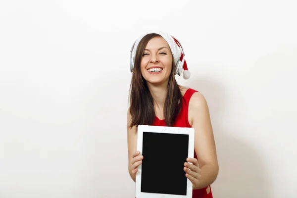 Mujer feliz bastante joven en vestido rojo y sombrero de Navidad escuchando música en los auriculares, la celebración de la tableta en el fondo blanco. Aparato de Santa niña aislado. Vacaciones de Año Nuevo 2018. Copiar anuncio de espacio . — Foto de Stock