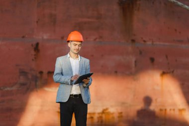 Yakışıklı tıraşsız başarılı iş adamı gri takım elbise, tablet, liman kargo paslı gemi arka plan ayakta tutan koruyucu inşaat turuncu kask. Erkek ile güneşli günde becerikli alet