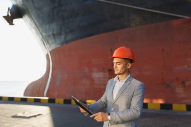Yakışıklı tıraşsız başarılı iş adamı gri takım elbise, tablet, liman kargo paslı gemi arka plan ayakta tutan koruyucu inşaat turuncu kask. Erkek ile güneşli günde becerikli alet