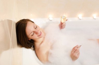 Uzun kahverengi saçlı, beyaz köpük küvet mum ışık banyoda yatan düz çıplak vücut çekici genç seksi kadın Şarap kadehi alkol ve sigara kapalı.