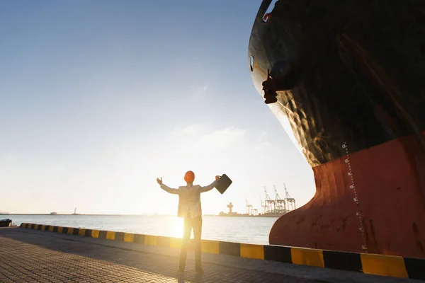 Young successful business man in gray suit and protective construction orange helmet holding case, spreading hands, standing in sea port against cargo rusty ship with water line, sun shine background