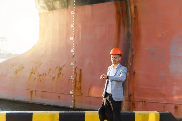 Handsome young successful business man in gray suit and protective construction orange helmet holding case, looking time on watch, standing in sea port against a cargo ship with water line background