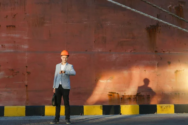 Handsome young unshaven successful business man in gray suit and protective construction orange helmet holding case, looking time on watch, standing in sea port against cargo rusty ship background
