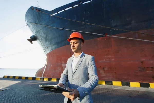 Handsome unshaven successful business man in gray suit, protective construction orange helmet holding tablet, standing in sea port against cargo rusty ship on water line background. Male with gadget