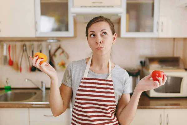 Eine junge verwirrte und nachdenkliche Frau in Schürze entscheidet sich für eine rote oder gelbe Tomate in der Küche. Diätkonzept. Gesunder Lebensstil. Kochen zu Hause. Essen zubereiten. — Stockfoto