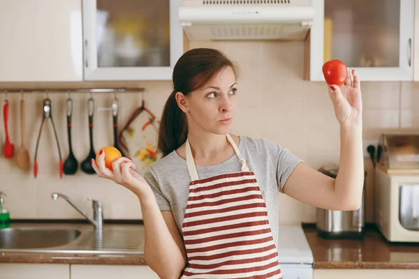 Молодая запутавшаяся и задумчивая женщина в фартуке решает выбрать красный или желтый помидор на кухне. Концепция диеты. Здоровый образ жизни. Готовить дома. Готовьте еду. . — стоковое фото