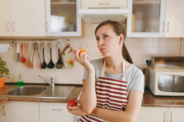 Eine junge verwirrte und nachdenkliche Frau in Schürze entscheidet sich für eine rote oder gelbe Tomate in der Küche. Diätkonzept. Gesunder Lebensstil. Kochen zu Hause. Essen zubereiten. — Stockfoto