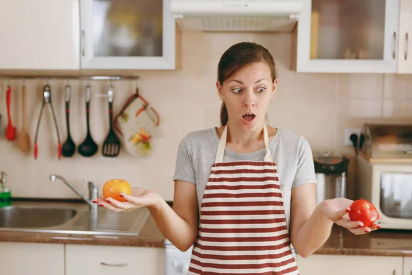 Молодая смущенная женщина в фартуке решает выбрать красный или желтый помидор на кухне. Концепция диеты. Здоровый образ жизни. Готовить дома. Готовьте еду. . — стоковое фото