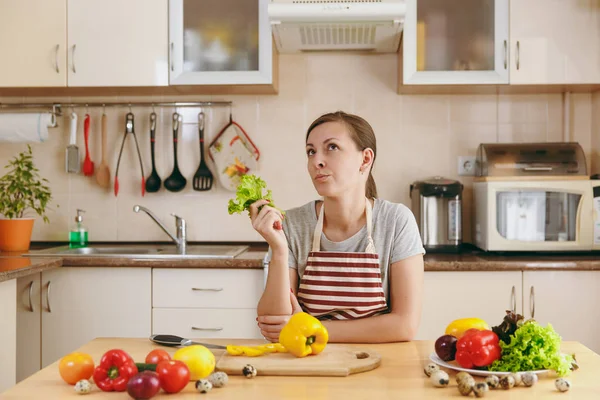 Una giovane donna attraente pensosa in un grembiule con foglia di lattuga in cucina. Il concetto di dieta. Uno stile di vita sano. Cucinare a casa. Preparare il cibo . — Foto Stock
