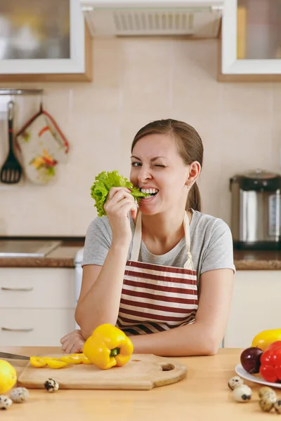 Молодая красивая женщина в фартуке с листком салата во рту на кухне. Концепция диеты. Здоровый образ жизни. Готовить дома. Готовьте еду. . — стоковое фото
