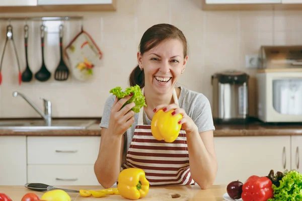 Молодая красивая женщина в фартуке с листьями салата и желтым перцем, смеявшаяся на кухне. Концепция диеты. Здоровый образ жизни. Готовить дома. Готовьте еду. . — стоковое фото
