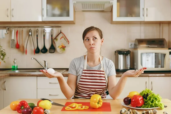 Una giovane donna attraente pensierosa in un grembiule sceglie tra uova di pollo e di quaglia in cucina. Il concetto di dieta. Uno stile di vita sano. Cucinare a casa. Preparare il cibo . — Foto Stock