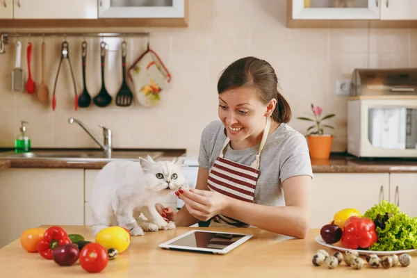 Μια νεαρή όμορφη γυναίκα με λευκή περσική γάτα στην κουζίνα με το tablet πάνω στο τραπέζι. Σαλάτα λαχανικών. Δίαιτα έννοια. Υγιεινού τρόπου ζωής. Το μαγείρεμα στο σπίτι. Προετοιμασία φαγητού. — Φωτογραφία Αρχείου