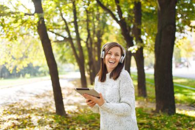 Güzel bir mutlu neşeli kahverengi saçlı Bayan Beyaz kazaklı bir tablet sonbahar Park'ta sıcak bir gün beyaz kulaklık müzik dinleme. Şehirde sonbahar.