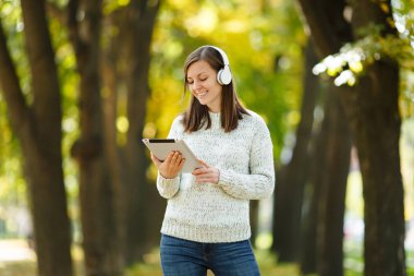 Güzel bir mutlu neşeli kahverengi saçlı Bayan Beyaz kazaklı bir tablet sonbahar Park'ta sıcak bir gün beyaz kulaklık müzik dinleme. Şehirde sonbahar.