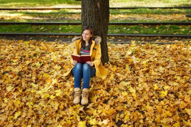 Güzel bir mutlu gülümseyen kahverengi saçlı kadın sarı ceket ve kot sıcak bir günde sonbahar Şehir Parkı kırmızı bir kitap ile akçaağaç ağacın altında oturuyor. Altın yaprak. Okuma kavramı