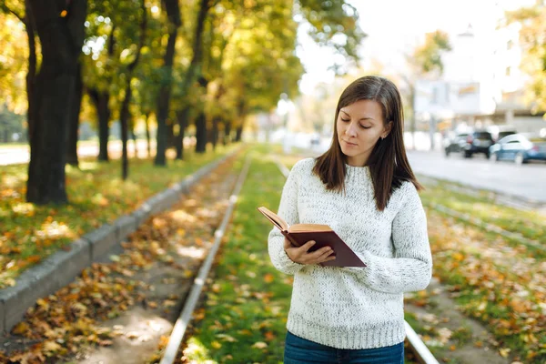 A beautiful happy smiling brown-haired woman in white sweater standing with a red book on the tram tracks in fall city park on a warm day. Autumn golden leaves. Reading concept.
