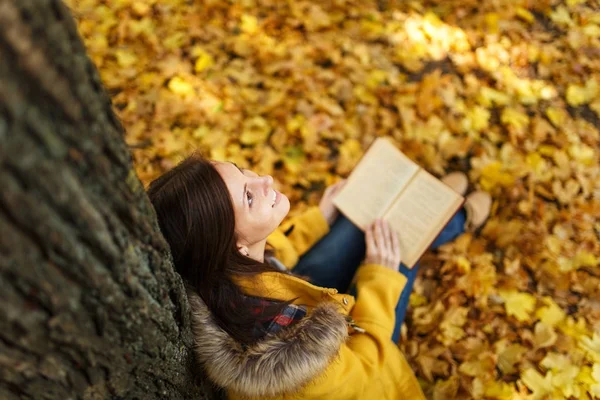 A beautiful happy smiling brown-haired woman in yellow coat and jeans sitting under the maple tree with a red book in fall city park on a warm day. Autumn golden leaves. Reading concept. Top view.