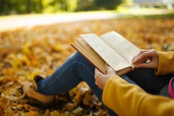 Kobieta w żółtym fartuchu i dżinsy, siedząc pod drzewem klonowym czerwony książkę w Upadek miasta park na ciepły dzień. Autumn golden odchodzi. Koncepcja czytania. Z bliska. — Zdjęcie stockowe