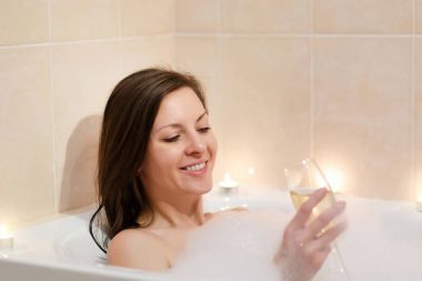 Uzun kahverengi saçlı ve düz çıplak vücut yalan, beyaz köpük küvet mum ışık banyoda, şarap cam kapalı içki alkol ile rahatlatıcı olan güzel seksi kadın.