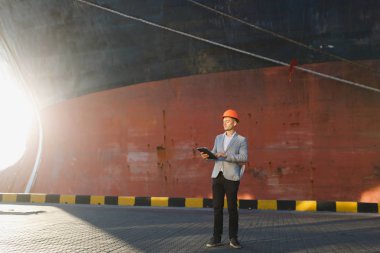 Yakışıklı tıraşsız başarılı iş adamı gri takım elbise, tablet, liman kargo paslı gemi arka plan ayakta tutan koruyucu inşaat turuncu kask. Erkek ile güneşli günde becerikli alet