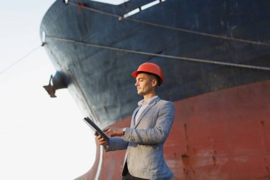 Yakışıklı tıraşsız başarılı iş adamı gri takım elbise, tablet, liman kargo paslı gemi arka plan ayakta tutan koruyucu inşaat turuncu kask. Erkek ile güneşli günde becerikli alet