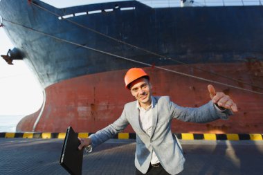 Yakışıklı mutlu başarılı iş adamı gri takım elbise, tablet holding, eller yayılan, liman kargo paslı geminin su hattı arka plan üzerinde karşı ayakta koruyucu inşaat turuncu kask