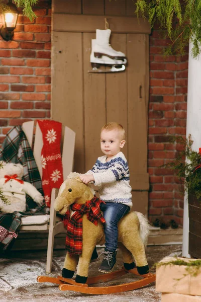 Παιχνιδιάρικο χαμογελά ευτυχισμένη χαριτωμένο μικρό παιδί αγόρι ντυμένο με πουλόβερ και τζιν που κάθεται στο κουνιστό άλογο στο διακοσμημένο δωμάτιο στο σπίτι με το νέο έτος. Χριστούγεννα καλή διάθεση. Τρόπου ζωής, οικογένεια και διακοπές έννοια 2018 — Φωτογραφία Αρχείου